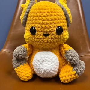 May include: A crocheted plush toy in the shape of a yellow and gray creature with large black eyes. The toy is sitting on a brown leather chair.