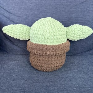 May include: A crocheted plush toy shaped like a character from the Star Wars franchise. The toy is green with brown ears and a brown base.