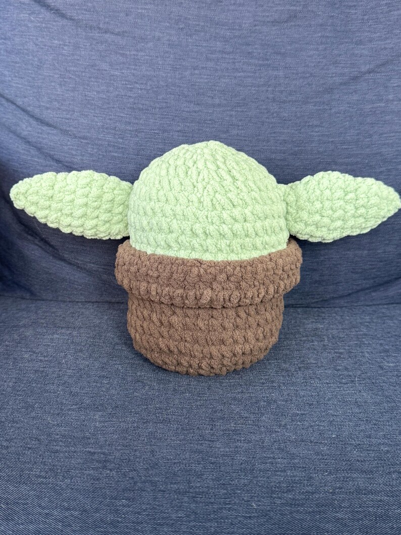 May include: A crocheted plush toy shaped like a character from the Star Wars franchise. The toy is green with brown ears and a brown base.