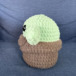 May include: A crocheted plush toy of a green character with large eyes, sitting in a brown crocheted pot.