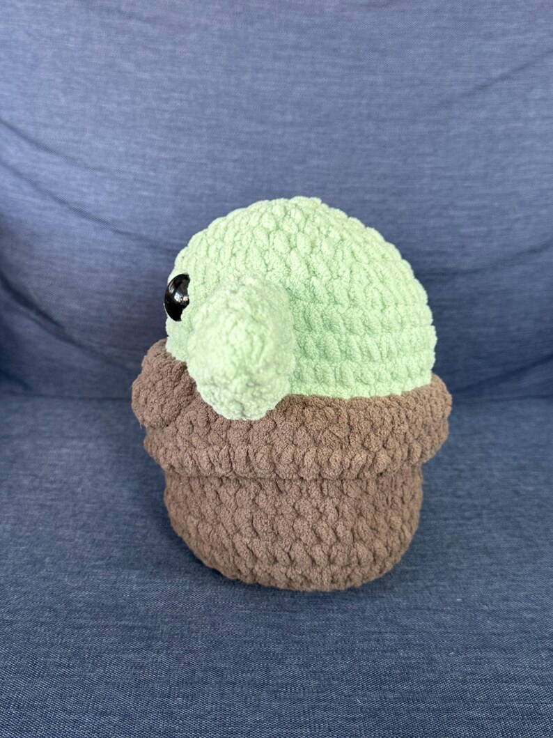 May include: A crocheted plush toy of a green character with large eyes, sitting in a brown crocheted pot.