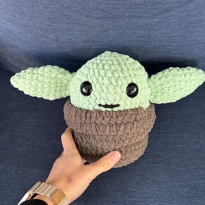 May include: A green crocheted plush toy with large black eyes and a brown crocheted collar. The toy is shaped like a small creature with large ears.