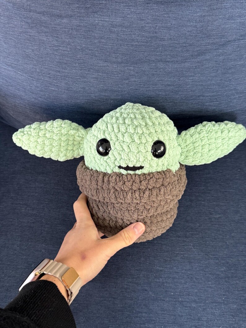 May include: A green crocheted plush toy with large black eyes and a brown crocheted collar. The toy is shaped like a small creature with large ears.