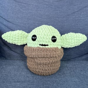 May include: A crocheted plush toy of a green character with large black eyes and a brown crocheted robe. The toy is sitting on a blue fabric surface.