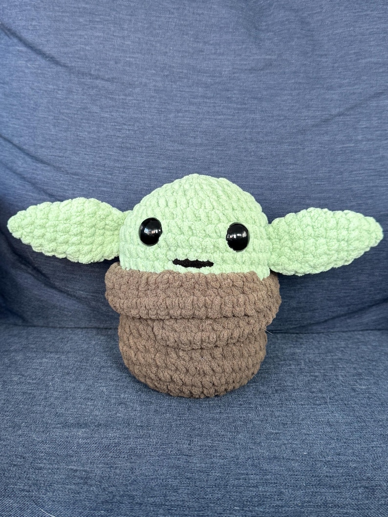 May include: A crocheted plush toy of a green character with large black eyes and a brown crocheted robe. The toy is sitting on a blue fabric surface.