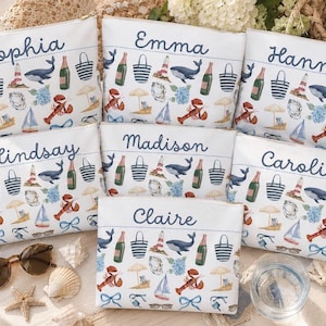 May include: Seven white rectangular zipped pouches, each personalised with a name in blue script. The fabric features a repeating nautical pattern including whales, lobsters, lighthouses, champagne bottles, beach bags, sailboats, and blue bows.