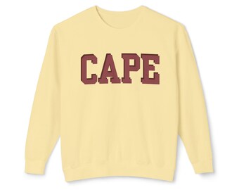 Könnte beinhalten: Hellgelbes Sweatshirt mit dem Wort "CAPE" in kastanienbraunen Blockbuchstaben auf der Brust. Das Sweatshirt hat einen Rundhalsausschnitt und lange Ärmel. Der Stoff scheint weich und bequem zu sein.