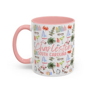 May include: White ceramic coffee mug with a pink rim. The mug features a colourful pattern of Charleston, South Carolina landmarks, including a pineapple fountain, a sailboat, and a crab. The text "Charleston, South Carolina" is printed in pink on the mug.