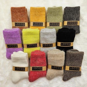May include: A collection of folded socks in various colors, including brown, yellow, green, gray, purple, and red. Each pair is wrapped with a band that reads "LOCOS". The socks are displayed on a white, fluffy surface.