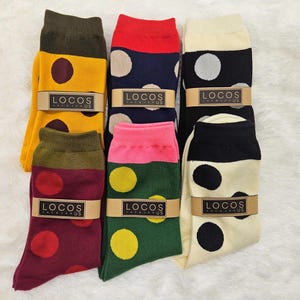 Puede incluir: Seis pares de calcetines coloridos con lunares. Los calcetines presentan varias combinaciones de colores, incluyendo amarillo con lunares marrones, azul marino con lunares beige y negro con lunares blancos. Cada par está envuelto con una banda de papel marrón que dice "LOCOS".