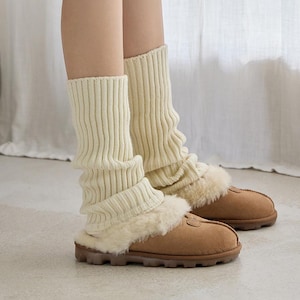 May include: Cream-colored knit leg warmers with a ribbed design, paired with brown suede slippers featuring a fluffy, light-colored trim. The leg warmers are scrunched down, creating a layered look.