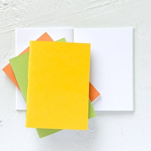 May include: A stack of three notebooks with solid-colored covers in bright yellow, lime green, and orange, layered over an open notebook with blank white pages. The notebooks are rectangular with rounded corners and a simple, clean design.