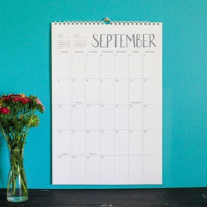 May include: A white wall calendar for September hangs against a teal wall. The word "September" is written in a handwritten font. A vase of flowers sits on a black surface in front of the wall.