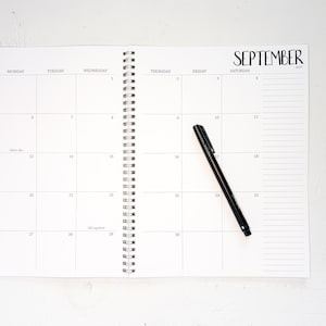 May include: A spiral-bound monthly planner open to September 2027, featuring a grid layout with days of the week, numbered dates, and a lined notes column on the right. A black ink pen rests diagonally across the page on the 17th of the month.