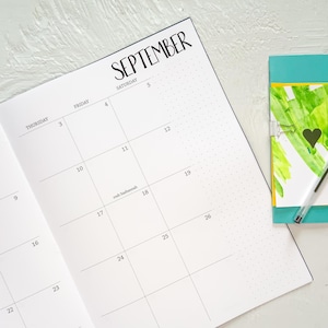 May include: Open planner page for September with the month's name in large, black lettering. The calendar grid is visible, with dates and days of the week. A small notebook with a green and yellow design and a black heart is on the right.
