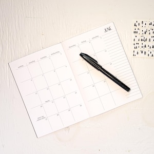 May include: An open planner showing a calendar for the month of June with black grid lines and text. The planner features days of the week, numbered dates, and specific labels like Flag Day and Father's Day. A black pen rests diagonally across the page.