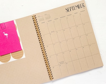 May include: A spiral-bound planner open to a September 2025 calendar page on tan paper with black grid lines and text. A bright pink rectangular note with a white flamingo graphic is clipped to the left side, which features a dot grid pattern.