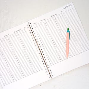 May include: An open spiral-bound weekly planner with white pages featuring black printed hourly schedules for Tuesday through Sunday, labeled July 25-31. A light peach-colored pen with a green and blue cap rests on the right page.