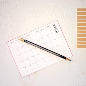 May include: A white paper calendar page for the month of August with black grid lines and text, featuring a black wooden pencil with a gold ferrule and white eraser resting diagonally across the center of the calendar grid.