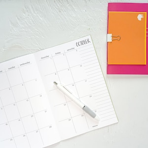 May include: Open notebook with an October calendar page and a white pen with a gray grip. The calendar is open to the month of October, with dates and lines for notes. A pink notebook with an orange page is clipped to the side.