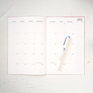 May include: Open notebook calendar for April 2027 with a white and blue Mildliner highlighter pen. The calendar is open to a blank spread with the days of the week across the top. The notebook has a pink cover.