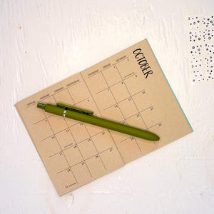 May include: A tan paper calendar for the month of October with black grid lines and text, featuring a matte olive green retractable ballpoint pen resting diagonally across the page. A small square sheet with black circular patterns is visible.