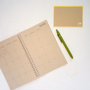 May include: A spiral-bound planner open to an August calendar page with tan paper and black text, a matching tan rectangular card with a yellow border and a small white mountain icon, and a solid olive green retractable ballpoint pen.
