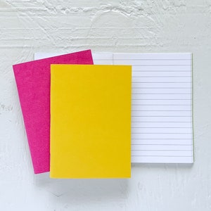 May include: Two notebooks, one with a bright yellow cover and the other with a bright pink cover. The yellow notebook is open to reveal lined pages.