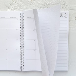 May include: A white spiral-bound planner with the month of July visible. The open pages display a calendar layout with numbered dates and lined pages. The planner is designed for organization and note-taking.