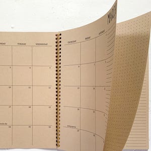 May include: A spiral-bound planner with tan pages featuring a monthly calendar layout for March. The grid includes days of the week, numbered dates, and text for St. Patrick's Day and the spring equinox. The right page has a dot grid pattern.