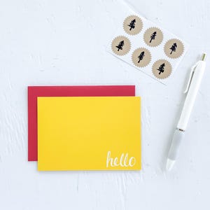 May include: A yellow card with the word "hello" in white script, layered over a red card. A sheet of brown stickers with black tree designs and a white pen with a gray grip are also present. The items are on a white surface.