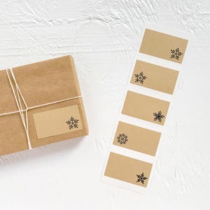 May include: A wrapped brown paper package with a blank label and a sheet of matching labels. Each label features a black snowflake design, perfect for holiday gift wrapping. The package is tied with twine.