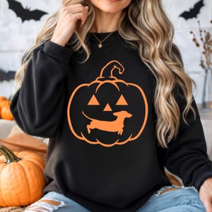 May include: Black sweatshirt with an orange pumpkin design. The pumpkin has a dachshund dog silhouette inside, creating a jack-o'-lantern face.