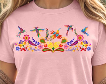 May include: A pink t-shirt with a colorful embroidered design of hummingbirds and flowers. The design features a variety of colors, including red, orange, yellow, green, blue, and purple.