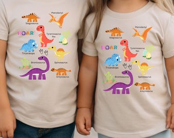 Könnte beinhalten: Zwei beige T-Shirts mit Dinosaurier-Motiv. Die Shirts zeigen bunte Illustrationen verschiedener Dinosaurier, darunter ein Stegosaurus, ein Tyrannosaurus Rex, ein Triceratops, ein Brontosaurus, ein Spinosaurus und ein Ankylosaurus. Der Text "ROAR!" ist auf beiden Shirts aufgedruckt.