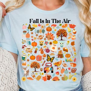 May include: Light blue t-shirt with the text "Fall is in the air" and a colourful graphic of autumn-themed icons, including pumpkins, leaves, owls, and a turkey.