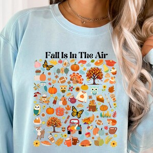 以下が含まれることがあります： 薄い青色のスウェットシャツに、秋のテーマのアイコンと「Fall is in the Air」というテキストがプリントされています。