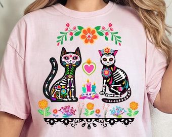Könnte beinhalten: Pinkes T-Shirt mit einem Design zum Tag der Toten, das zwei schwarze Katzen mit bunten Zucker Schädel Gesichtern und floralen Akzenten zeigt. Die Katzen sind von Blumen und Kerzen umgeben.