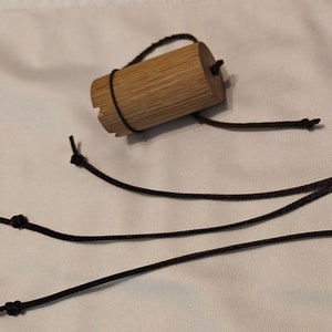 May include: A small wooden cylinder with a brown leather cord attached. The cord is tied in knots at the ends and is long enough to be worn as a necklace.