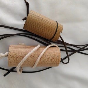 Könnte beinhalten: Zwei zylindrische Holzobjekte mit braunen Lederschnüren und weißen Seilakzenten. Ein Objekt hat ein schwarzes Band. Die Objekte sind hellbraun und scheinen handgefertigt zu sein. Der Hintergrund ist ein weißer Stoff.