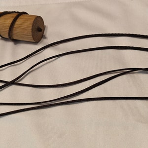 May include: A small, cylindrical wooden container with a dark brown cord wrapped around it, alongside several long, dark brown cords with knots at the ends. The items are on a white surface.
