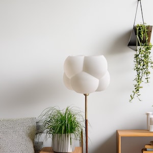 Peut inclure: Une lampe blanche en forme de nuage avec un pied doré est posée sur une table d'appoint en bois à côté d'un fauteuil gris. Une plante en pot est posée sur la table. Une plante suspendue avec des feuilles vertes pend du mur.