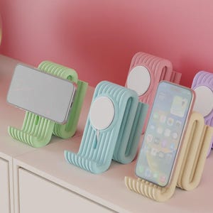 Può includere: Una collezione di supporti per telefono colorati in tonalità pastello di verde, blu, rosa, giallo e viola. Ogni supporto ha un design unico con una forma curva e un elemento bianco circolare. Sono esposti degli smartphone.