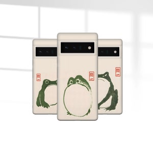 May include: Three phone cases with a beige background and a green frog design. The frog is in a circle with the Chinese character "関" in red on the top right corner of each case.