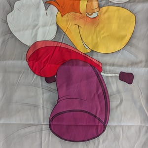 May include: A cartoon character pillowcase featuring a yellow-faced character with orange hair, a red hat, and a purple body. The character is lying on a gray background with white accents. The design is a vibrant and playful.