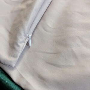 May include: Close-up of a light gray satin fabric with a white zipper. The fabric is folded, revealing a glimpse of a dark green satin material underneath. The texture of the satin is smooth and reflective.