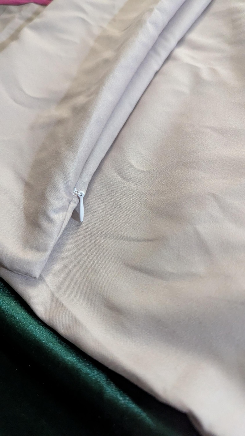 May include: Close-up of a light gray satin fabric with a white zipper. The fabric is folded, revealing a glimpse of a dark green satin material underneath. The texture of the satin is smooth and reflective.