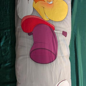 May include: A long, rectangular body pillow featuring a cartoon character with yellow hair, a red top, and a purple lower body. The pillow has a light gray background with cartoon elements.