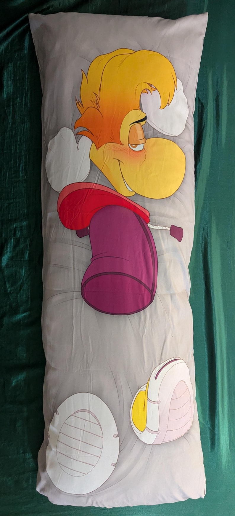 May include: A long, rectangular body pillow featuring a cartoon character with yellow hair, a red top, and a purple lower body. The pillow has a light gray background with cartoon elements.