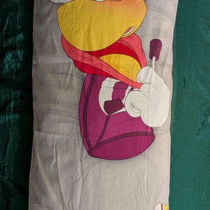May include: A long, gray body pillow featuring a cartoon character with yellow hair, a yellow beak, and a red scarf. The character's body is purple, and the pillow has a yellow and white design at the bottom.
